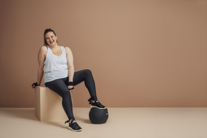 Imagem de uma mulher obesa, uma das condições para a síndrome metabólica. Ela está sentada em uma caixa e com o pé em cima de uma bola, usando roupa de ginástica, olhando para a câmera e sorrindo.