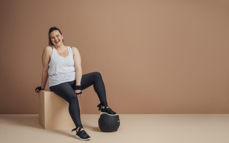 Imagem de uma mulher obesa, uma das condições para a síndrome metabólica. Ela está sentada em uma caixa e com o pé em cima de uma bola, usando roupa de ginástica, olhando para a câmera e sorrindo.