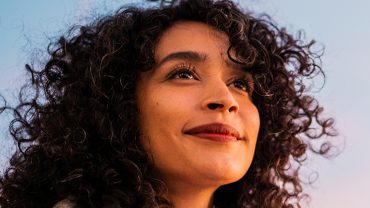 Mulher de cabelo cacheado olhando para o céu, com expressão de admiração e curiosidade.